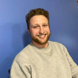 Picture of Philip Bard. He is 22 years old with curly brown hair and a beard, wearing a gray sweater against a dark blue background. He is smiling.